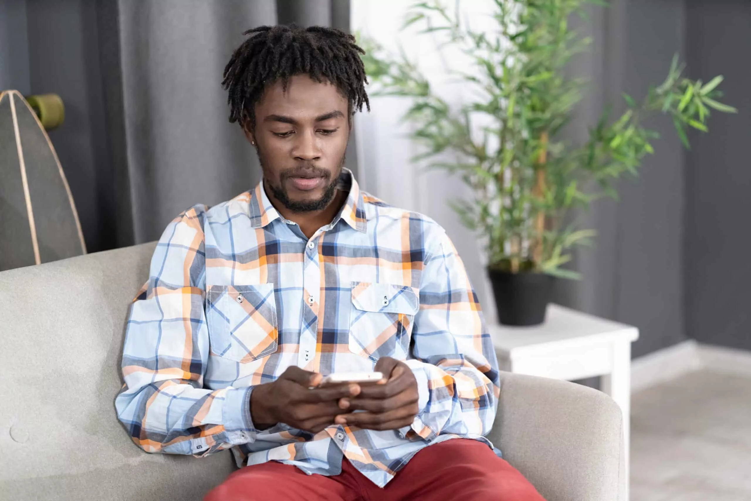 concentrated or interested african american man use smartphone checking social media or updating information. portrait of young man holding smartphone sitting on the sofa. social media concept